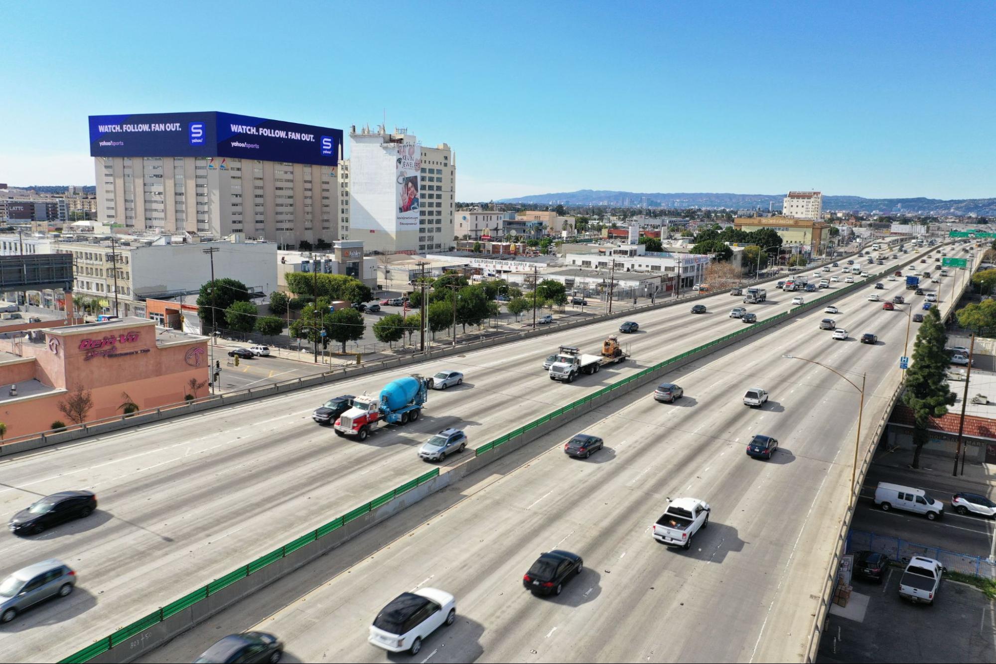 Aerial view of a busy freeway with cars and trucks, with large digital billboards on nearby buildings displaying 'Watch. Follow. Fan Out.' for Yahoo Sports.