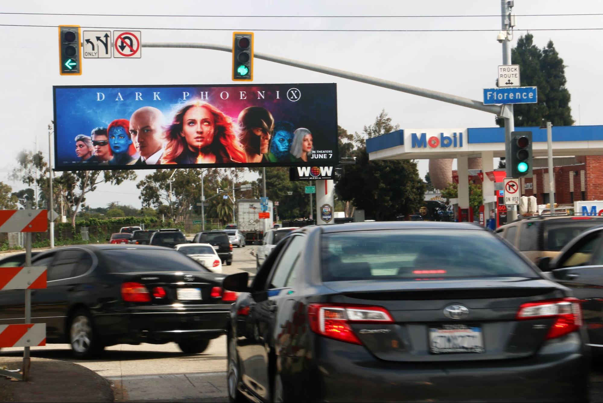 Street intersection with a digital billboard displaying the 'Dark Phoenix' movie poster featuring the main cast, located near a Mobil gas station."