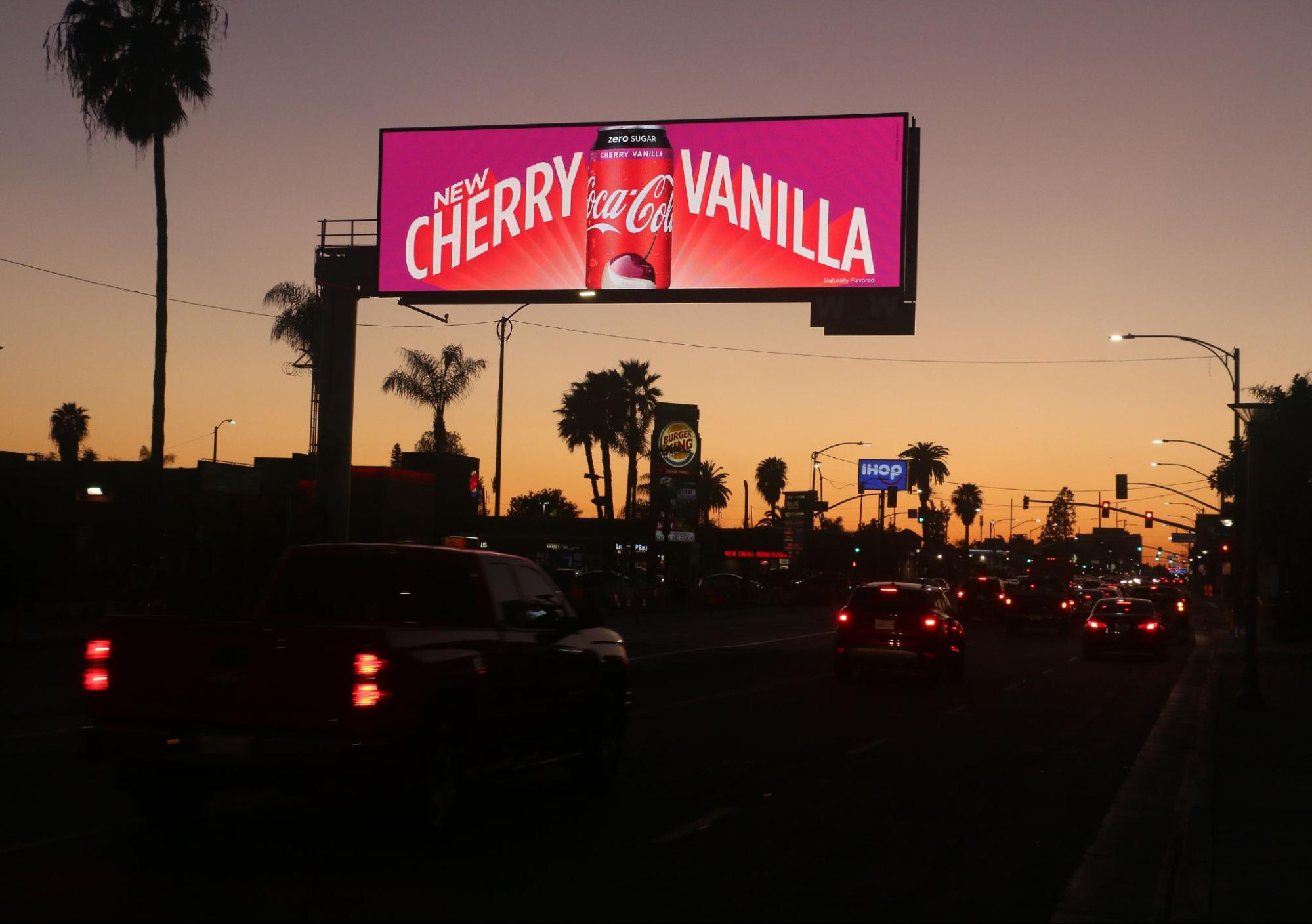 Dusk cityscape with a Coca-Cola Cherry Vanilla digital billboard, cars on the road, and palm trees in the background.