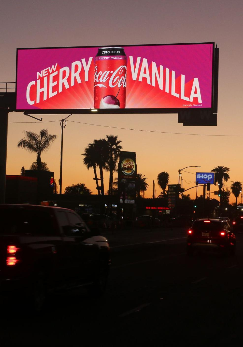City street at dusk with a large Coca-Cola Cherry Vanilla ad displayed on a digital billboard.