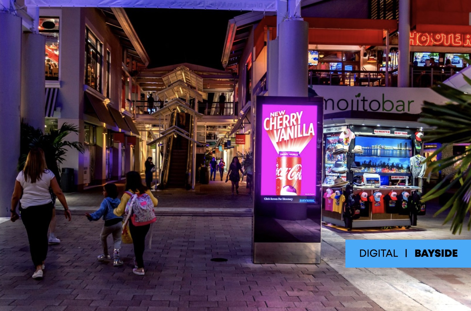 Night scene at Bayside shopping area with a digital billboard displaying Coca-Cola Cherry Vanilla ad, surrounded by shops and visitors.