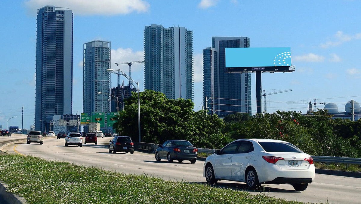 Urban road with vehicles driving toward tall skyscrapers, featuring an empty billboard space on the side.
