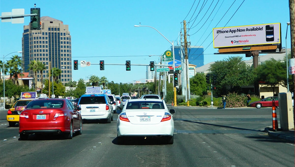Traffic at a Las Vegas intersection under a digital billboard advertising One Nevada’s iPhone app, with palm trees and office buildings in the background.
