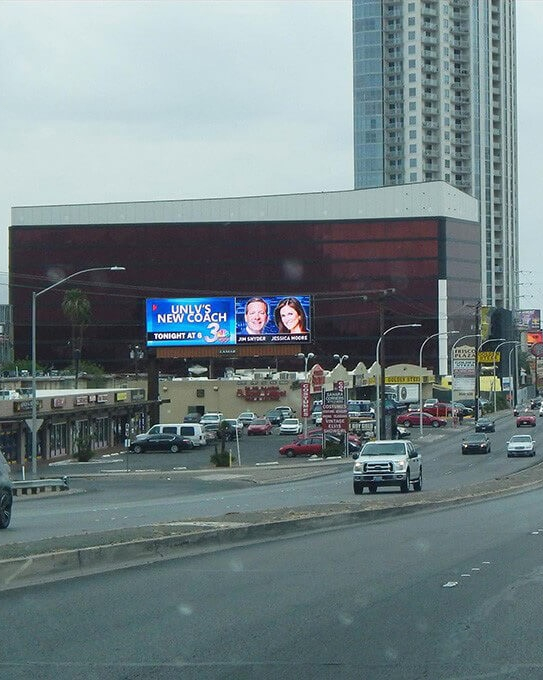 Digital billboard announcing UNLV’s new coach on a busy street, promoting an evening segment on Channel 3.